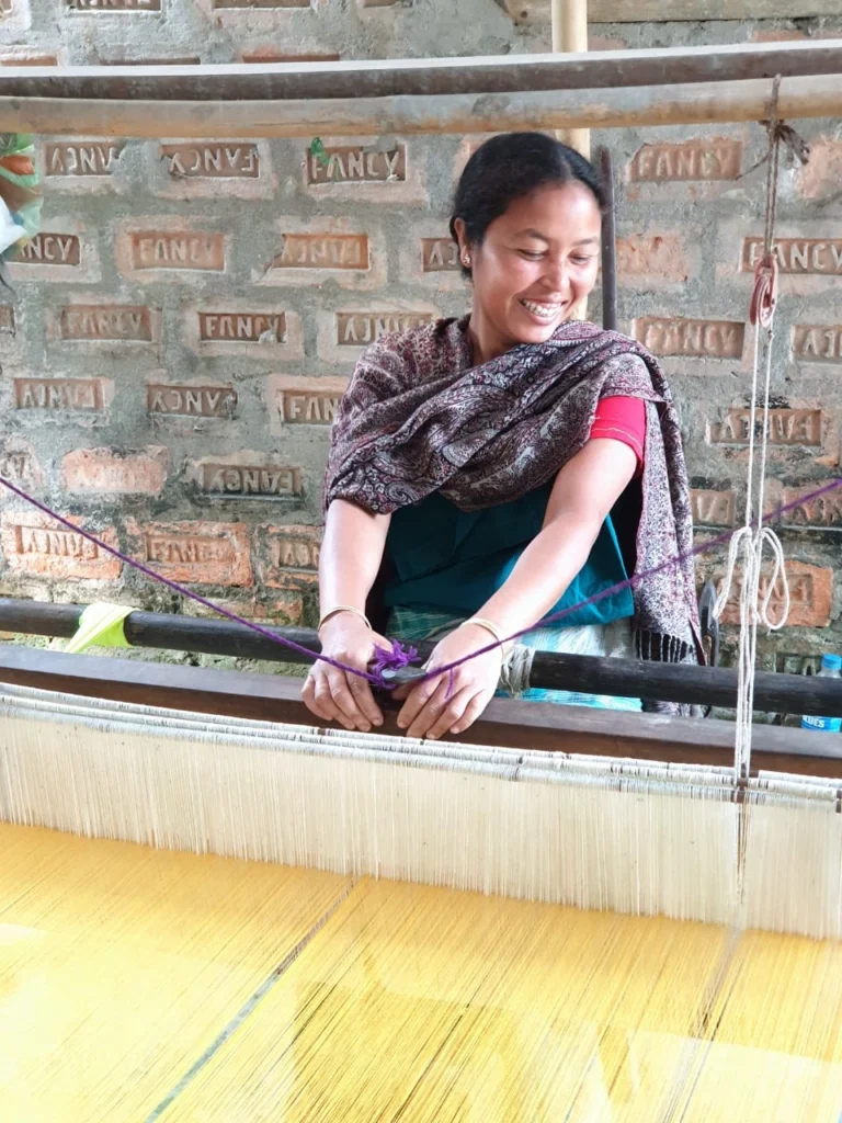 Assamese woman weaving on traditional handloom with silk threads in a rural setting