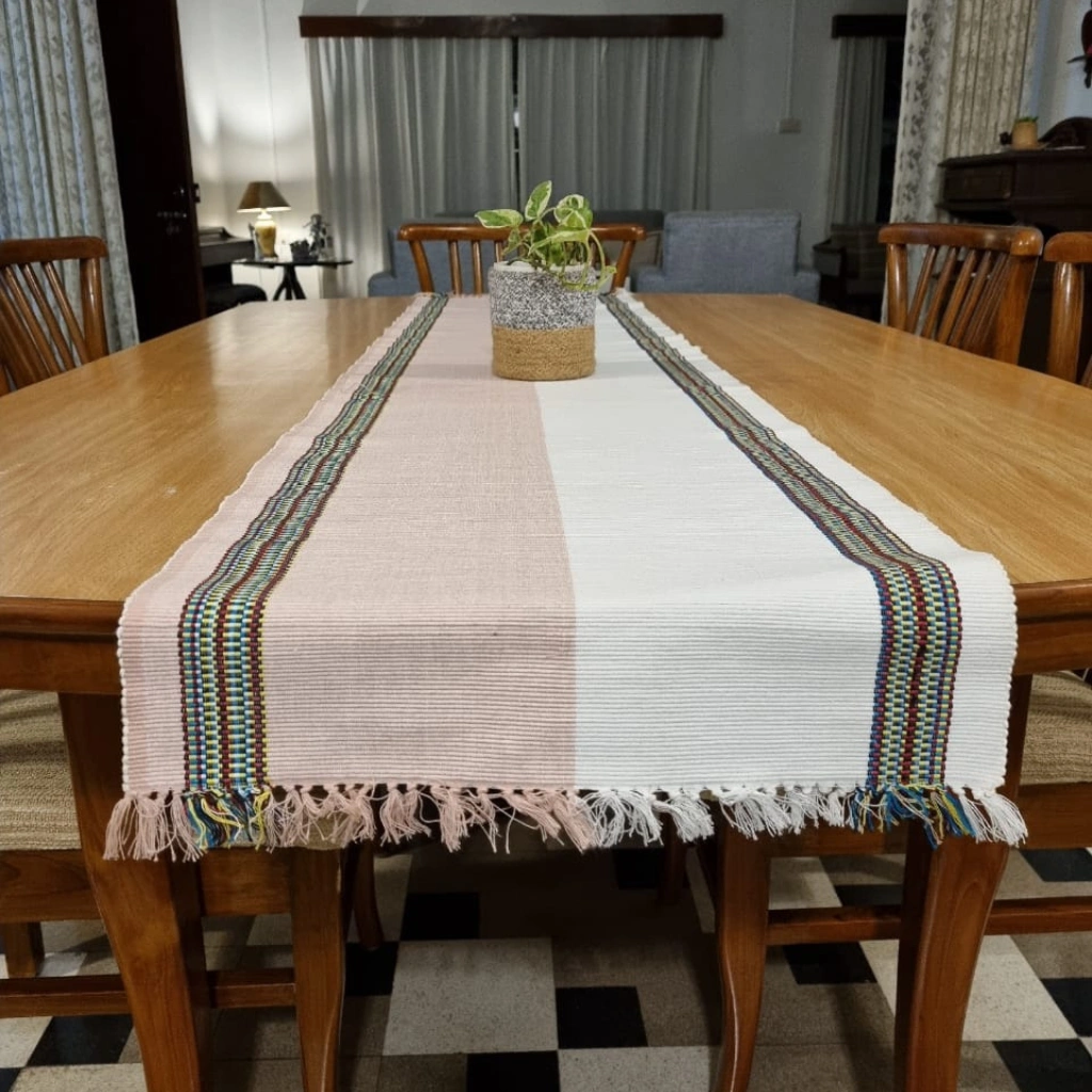 A long, handwoven cotton table runner with a white and blush pink base and colorful geometric borders, styled on a wooden dining table.