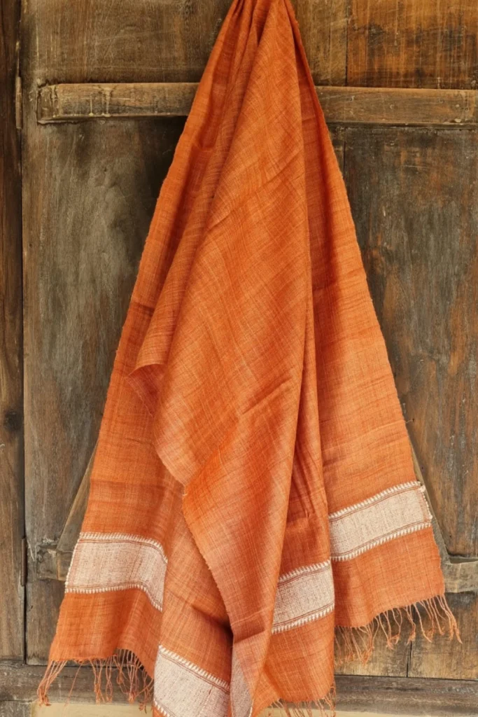Handwoven Ahimsa Eri silk stole in Indian madder dye draped against a rustic wooden background with soft fringes and woven border details.